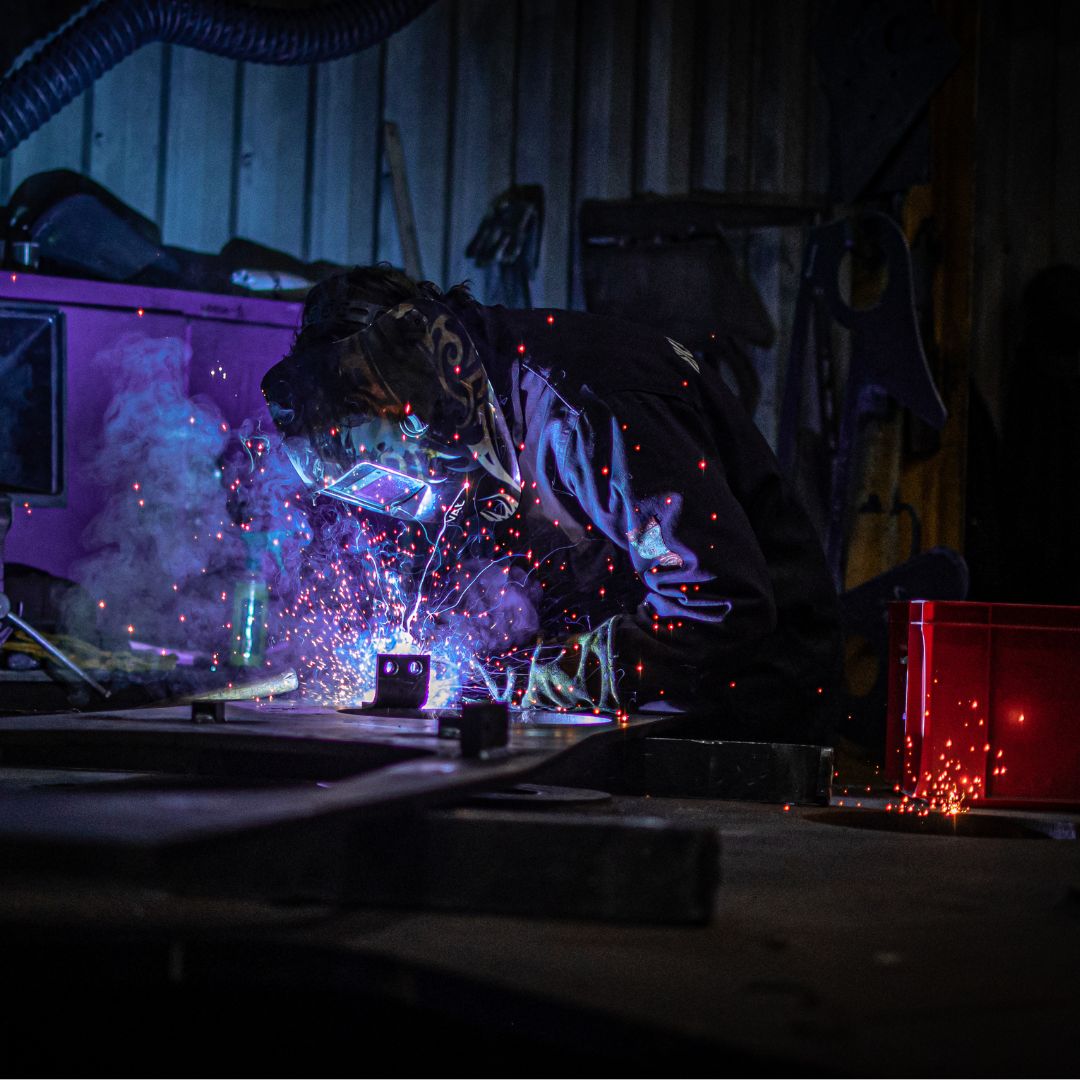 Person wearing welding mask and boiler suit welding a piece of metal with sparks flying.